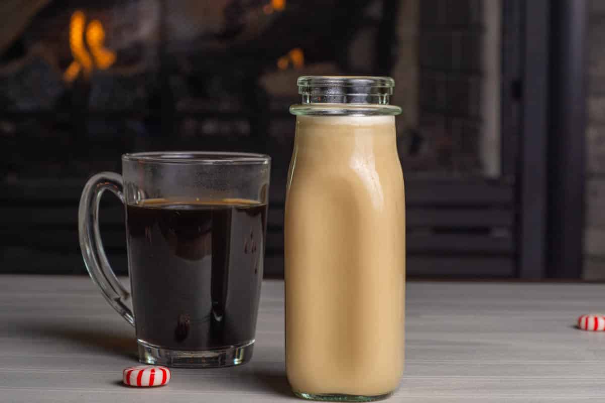 A clear mug of coffee with a small milk bottle filled with peppermint coffee creamer in front of a fireplace.