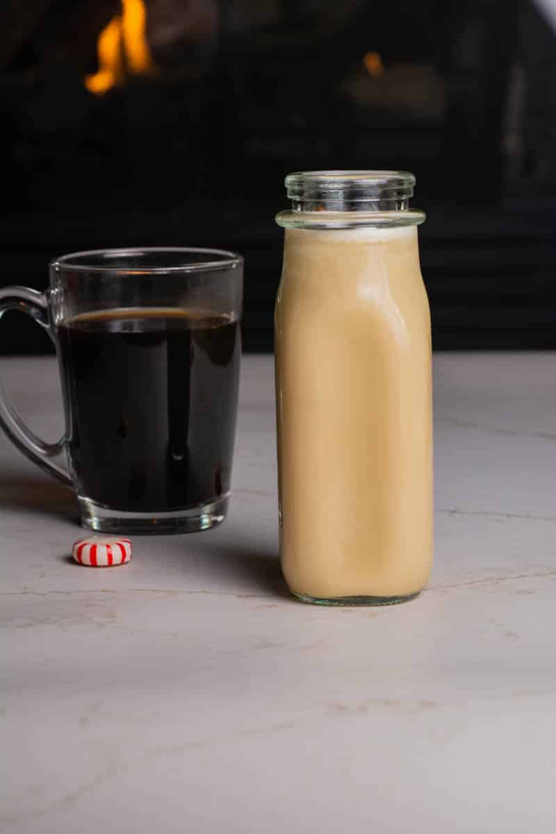 A clear mug of coffee with a small milk bottle filled with peppermint coffee cream in front of a fireplace.