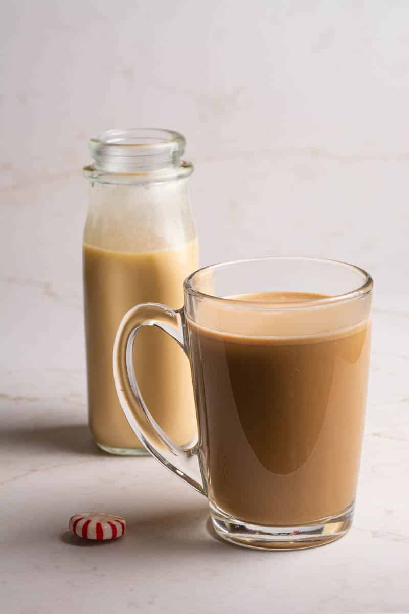 A clear mug of coffee with a small milk bottle filled with peppermint coffee cream and peppermint candies on a table.
