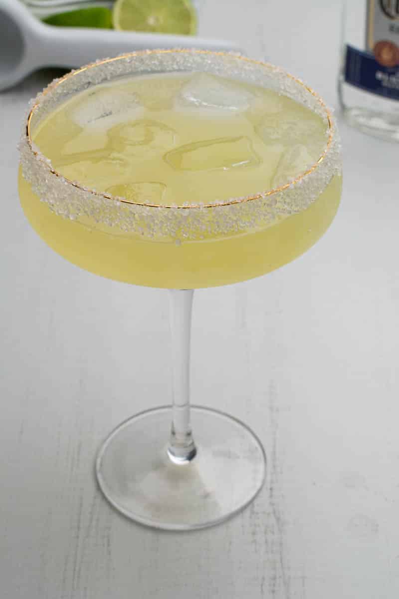 A close up photo of a margarita with agave. A citrus juicer and tequila is in the background.