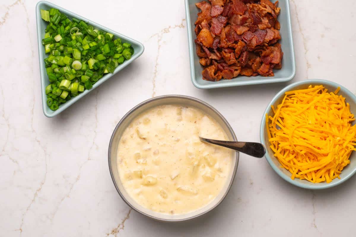 A bowl of potato soup with hashbrowns and three condiment bowl with fresh scallions, bacon, and shredded cheese.