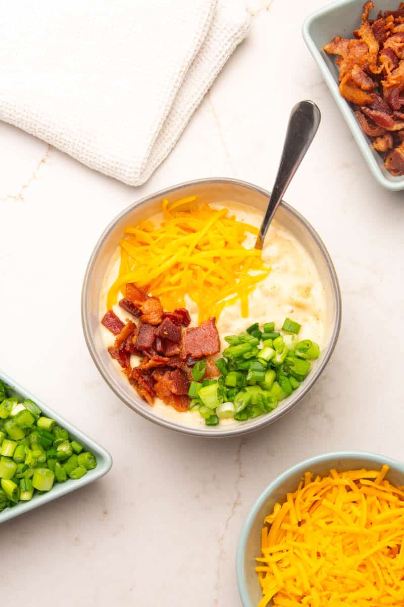 A round bowl of potato soup with hashbrowns. The hashbrown soup is garnished with bacon, shredded cheese and scallions.