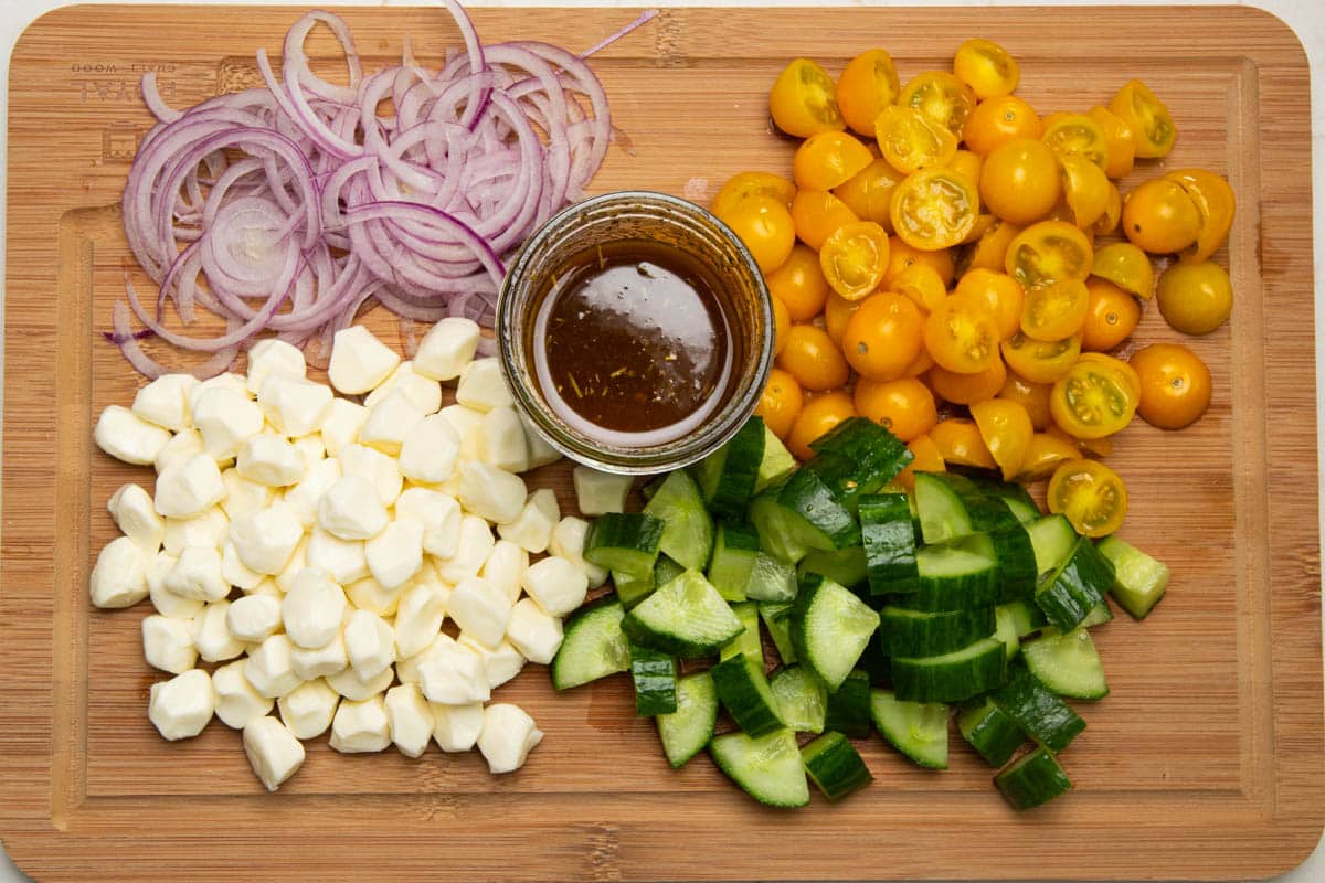 A wooden cutting board with sliced red onions, chopped grape tomatoes, chopped cucumbers, mozzarella pearls, and a balsamic vinaigrette dressing.