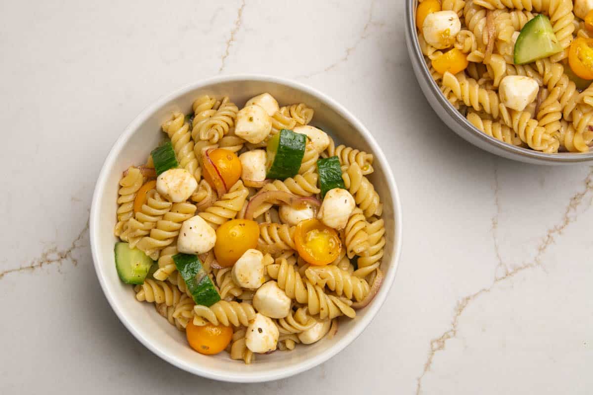 Two bowls of pasta with mozzarella pearls, yellow grape tomatoes, cucumbers, and sliced red onions.
