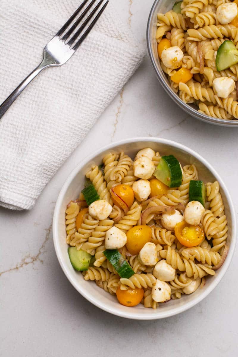 An overhead photo of a white bowl of pasta, mozzarella pears, grape tomatoes, cucumbers, and sliced red onions.