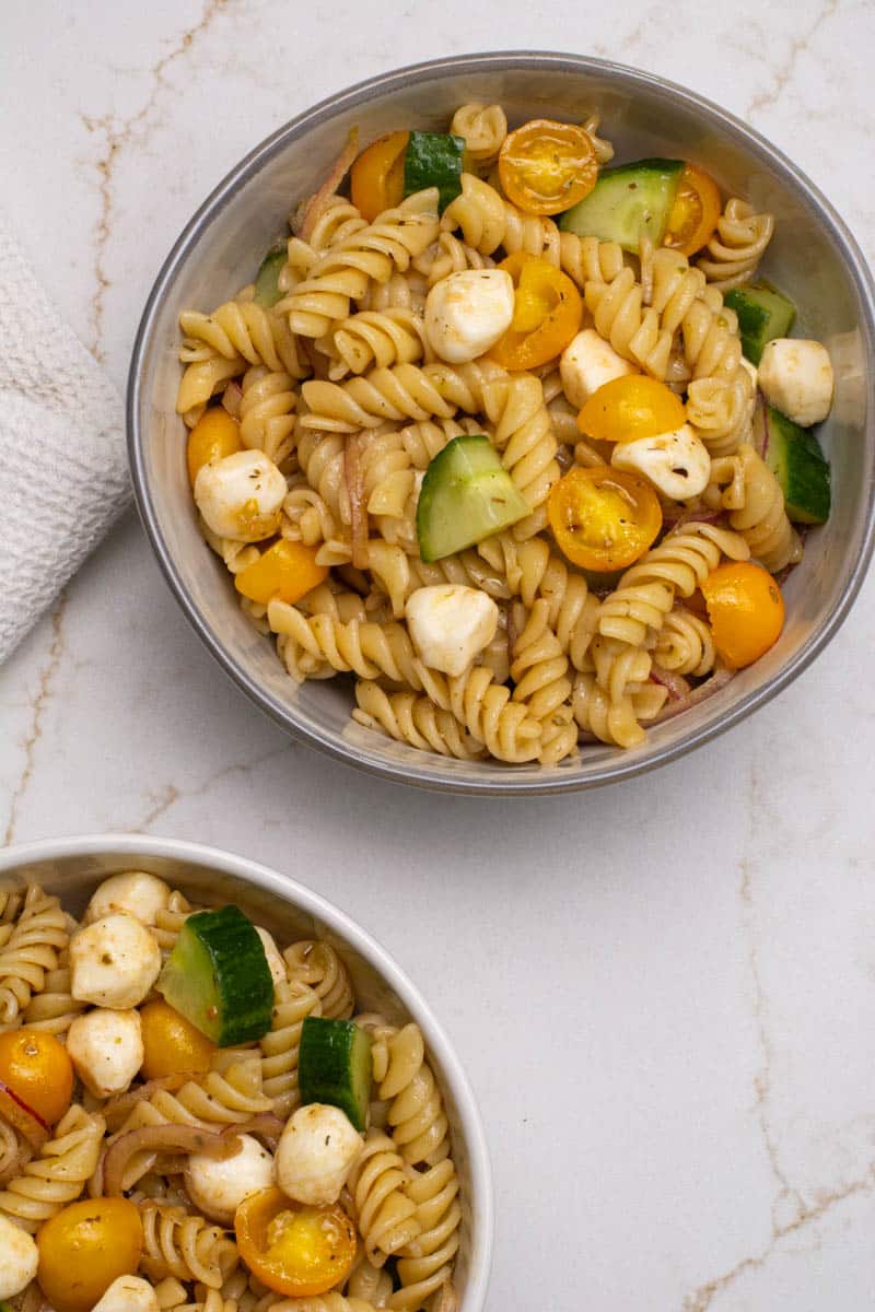 An overhead photo of two white bowls filled with mozzarella pasta salad with grape tomatoes, cucumbers and sliced onions.