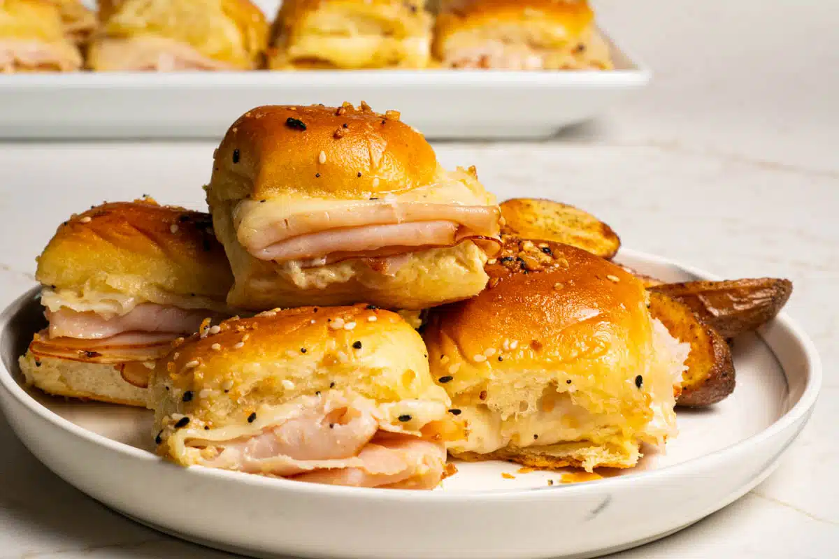 A stack of turkey sliders on a round white plate with additional sliders in the background.