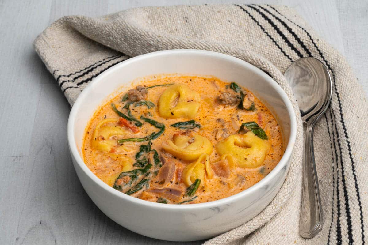 A white bowl of tortellini soup with sausage and spinach with a spoon and kitchen towel.