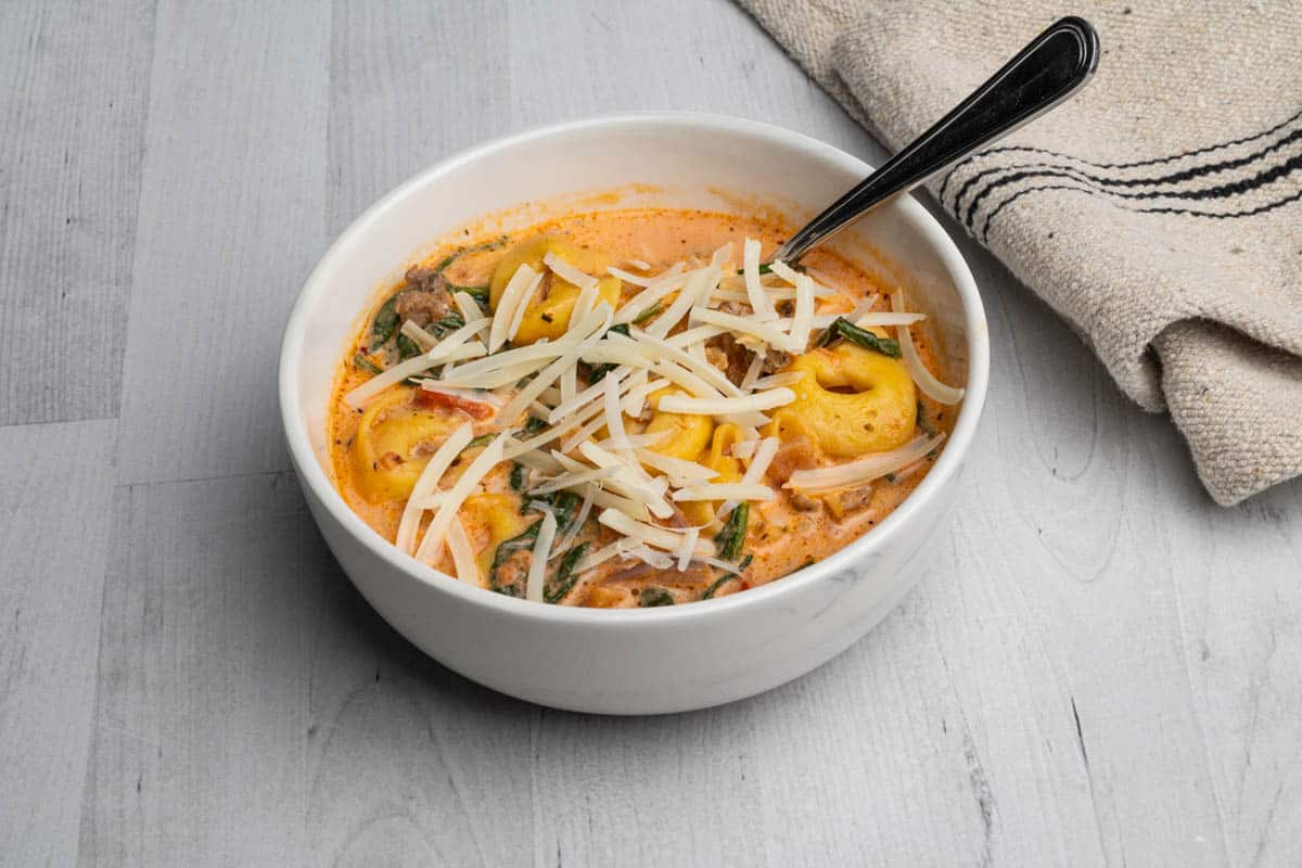 A white bowl of tortellini soup with sausage and spinach with a spoon and kitchen towel.