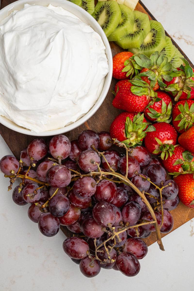 A wooden board on a table with a bowl of cool whip fruit dip, grapes, strawberries, and sliced kiwi.
