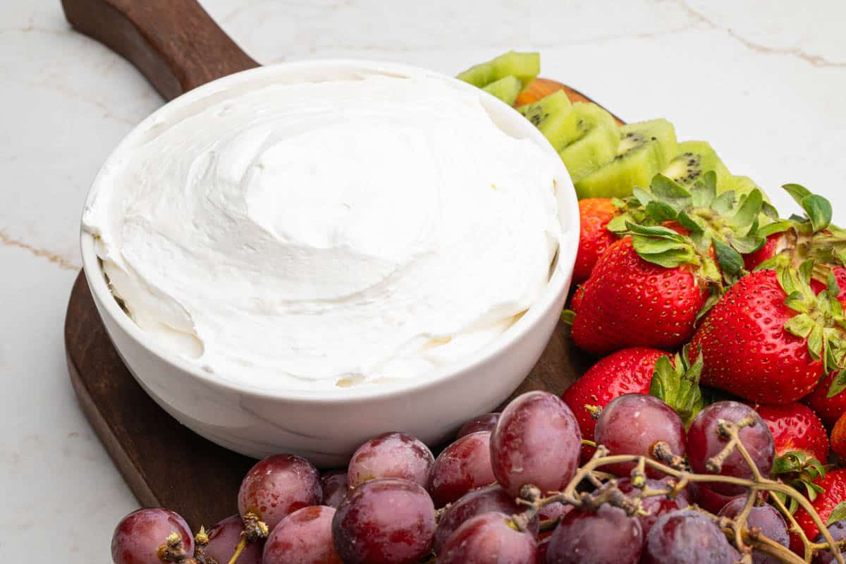 A wooden board on a table with a bowl of cool whip fruit dip, grapes, strawberries, and kiwi.
