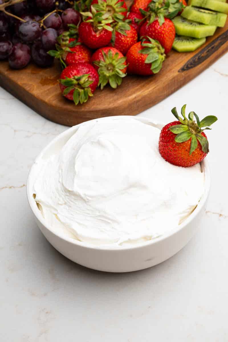 A wooden board on a table with a bowl of cool whip fruit dip, grapes, strawberries, and kiwi.