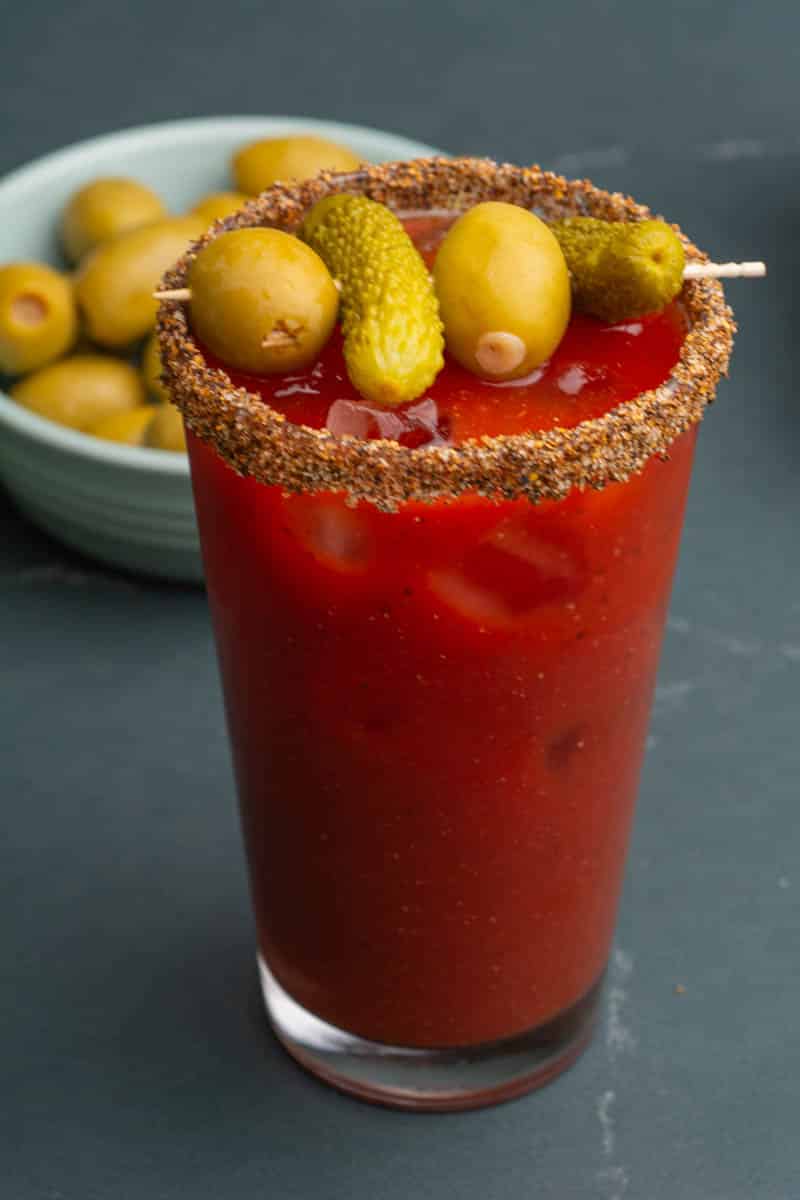 A virgin Bloody Mary mocktail in a pint glass with a small skewer of dill pickles and olives and olives in a bowl in the background.