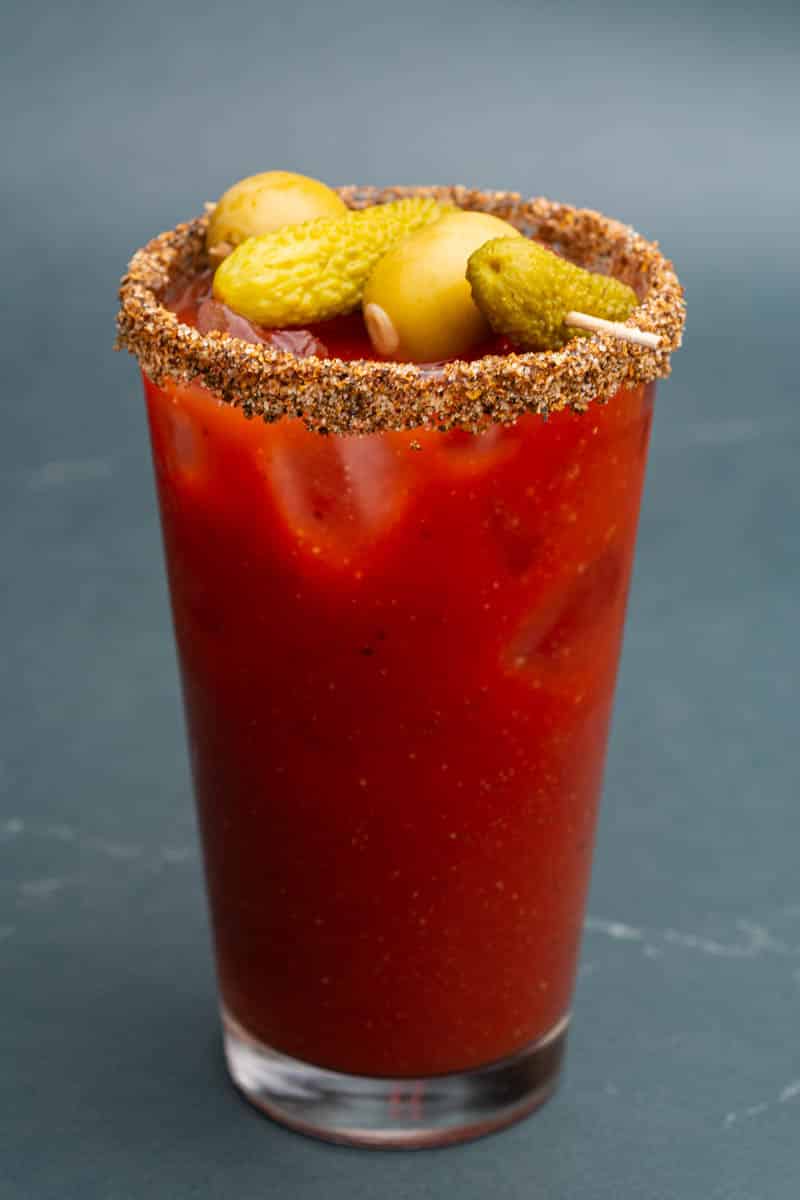A virgin Bloody Mary mocktail in a pint glass with a small skewer of dill pickles and olives.