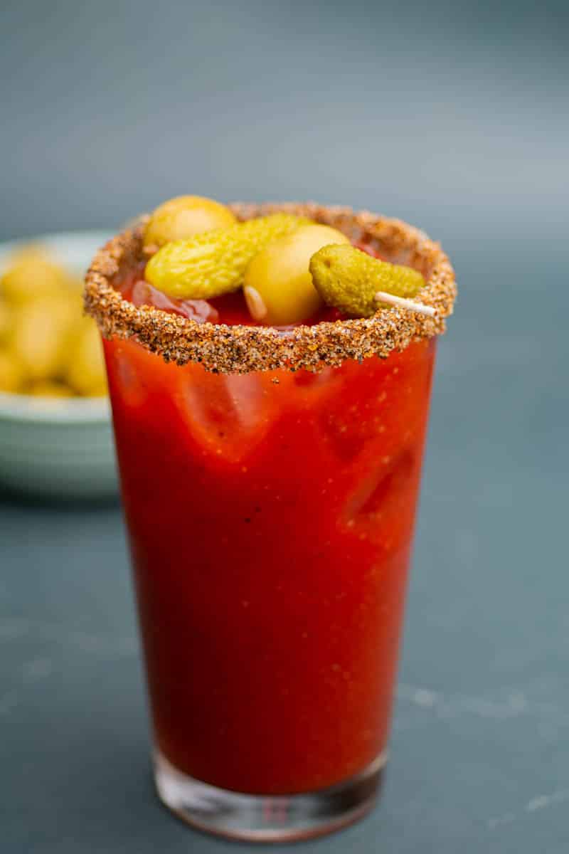 A virgin Bloody Mary drink in a tall pint glasss with a garnish skewer of pickles and olives and a bowl of olives in the background.