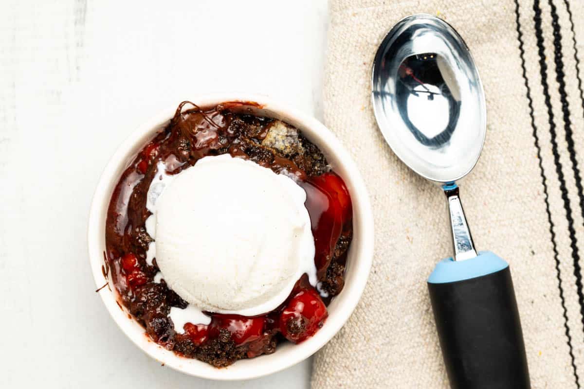 Chocolate cherry dump cake with ice cream scoop on it and a ice cream scoop and a kitchen towel.