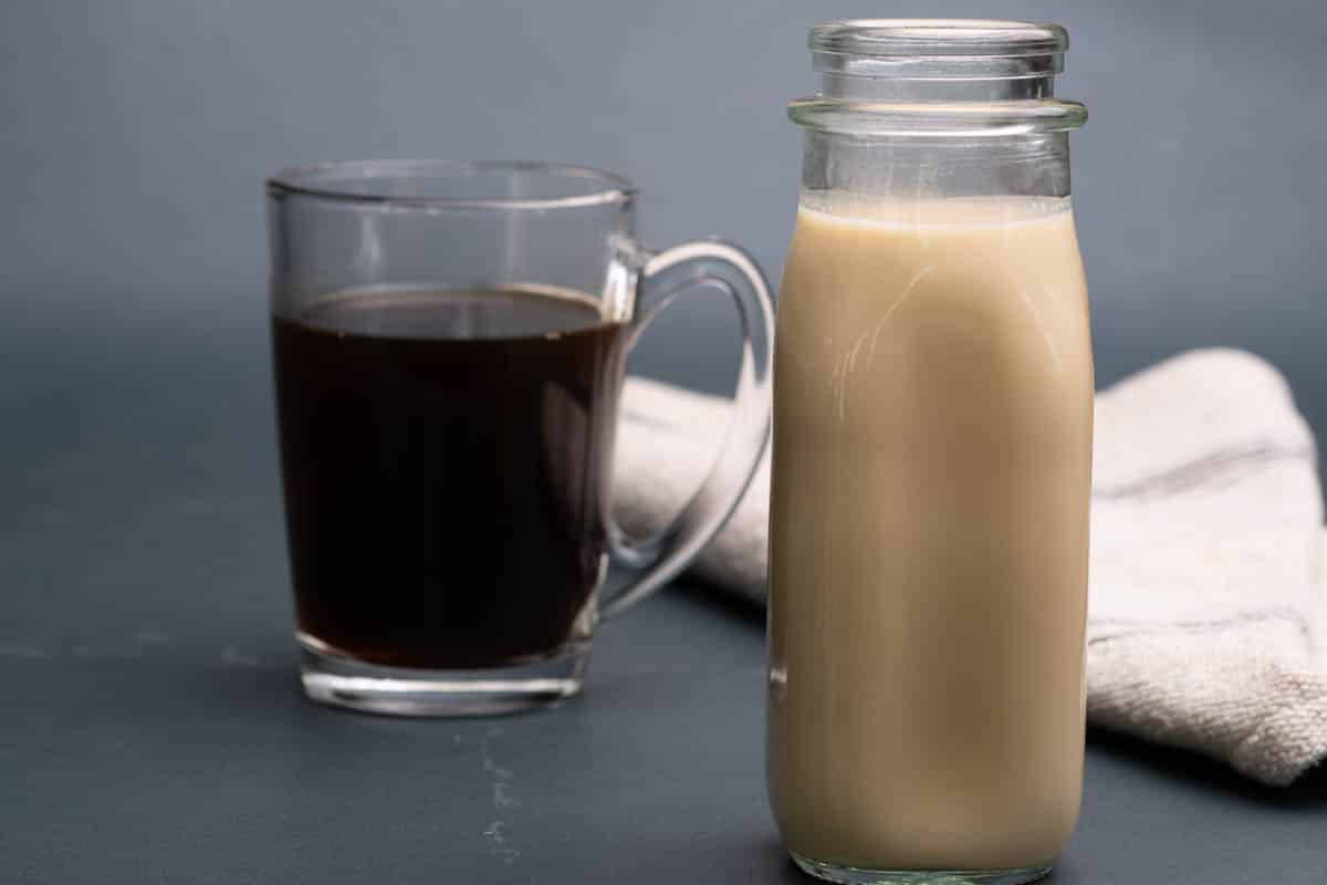 A glass cup of coffee with a jar of salted caramel coffee creamer in a glass jar with a kitchen towel in the background.