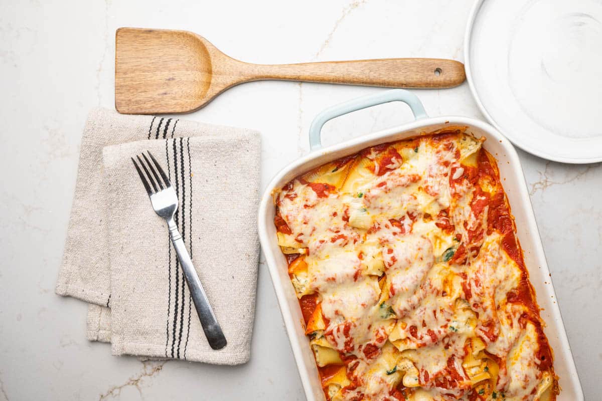 Overhead view of stuffed shell with ricotta cheese and spinach, a fork and kitchen towel, round plate, and wooden serving spatula.