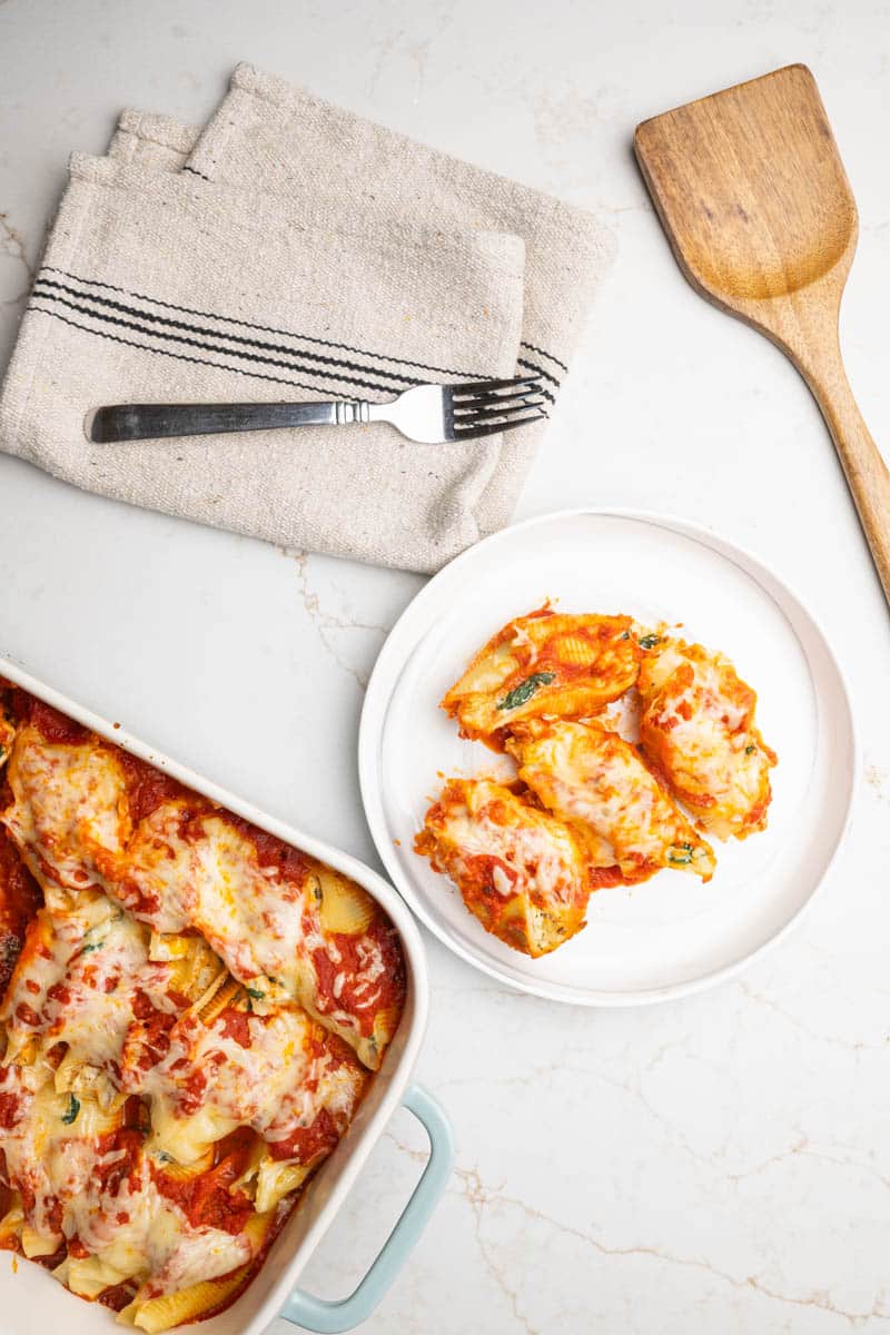 Overhead photo of stuffed shells with ricotta cheese and spinach in a rectangular baking dish, round plate, kitchen towel and serving spatula.