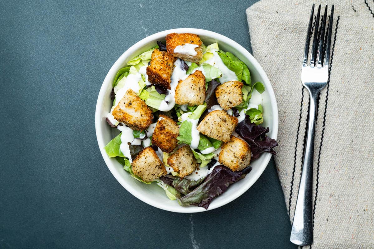 A bowl of air fryer croutons in a salad in a round bowl with a fork on a kitchen towel on a table.