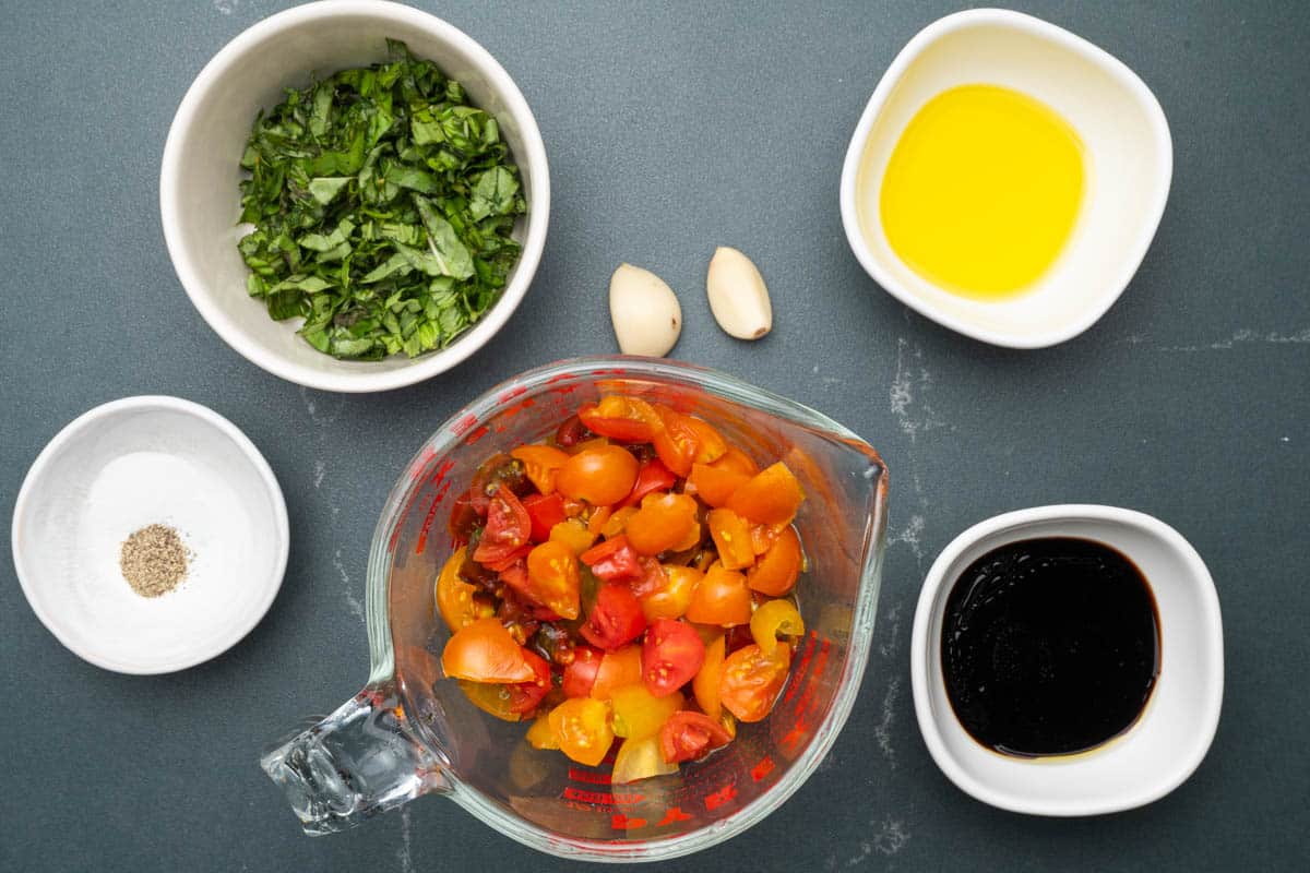 Overhead view of diced tomatoes, chopped basil, olive oil, balsamic glaze, salt, pepper, and garlic on a table.