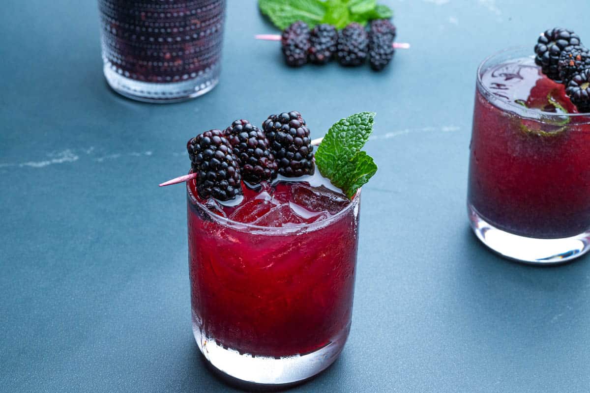 Two glasses of blackberry bourbon smash with a cocktail shaker and skewers of blackberries.