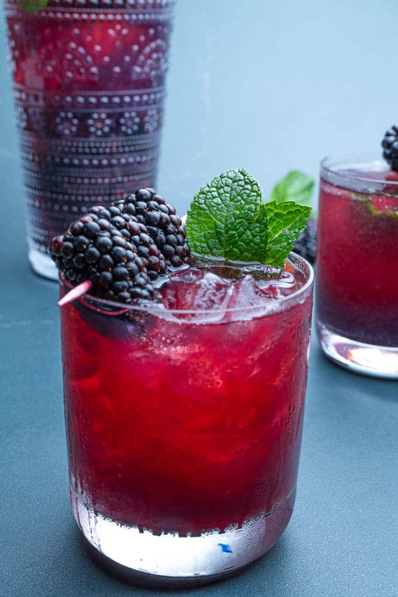 Blackberry bourbon smash in glasses with blackberry and mint garnishes, with a cocktail shaker in the background.