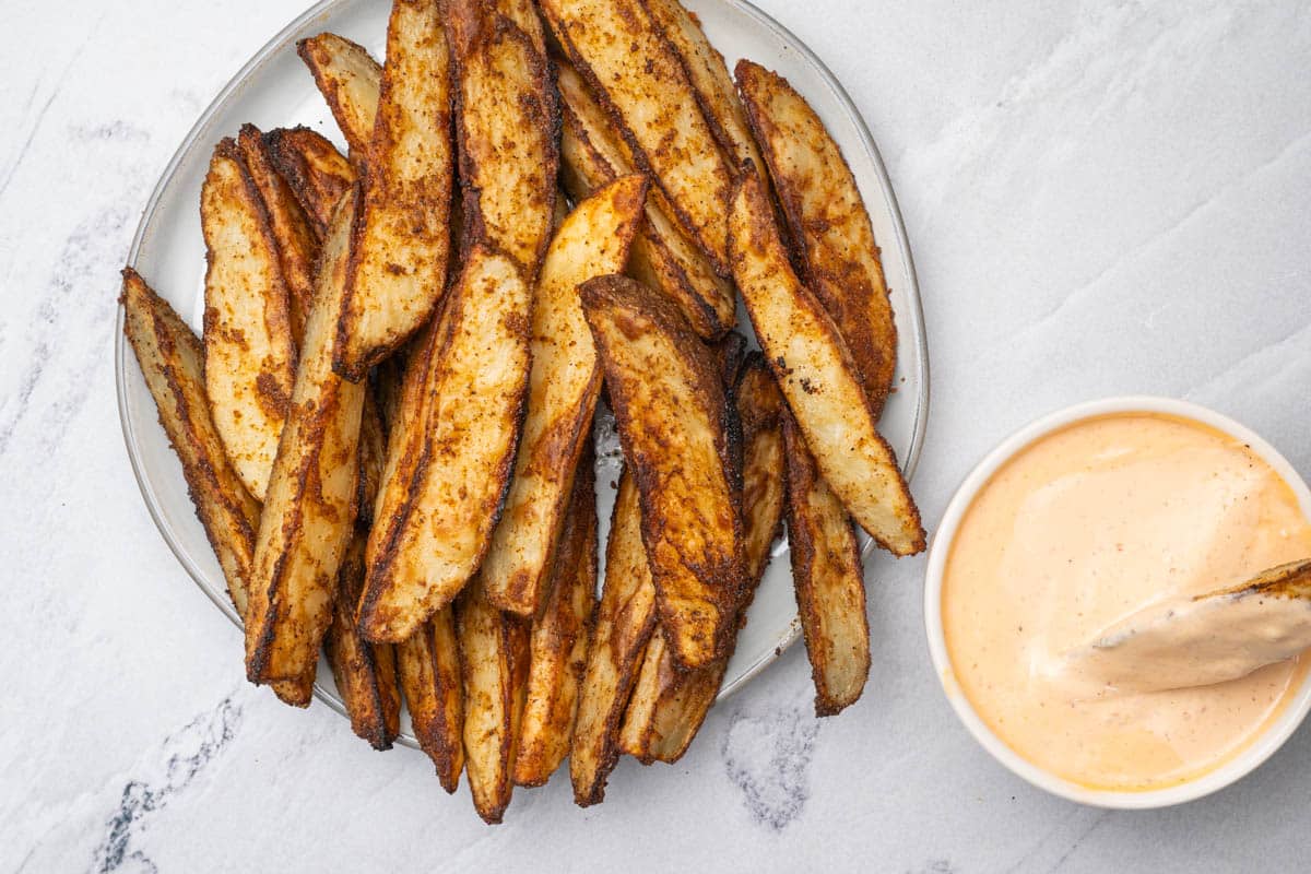 Oven roasted potato wedges on a round plate with a potato wedge dipped in sriracha aioli.