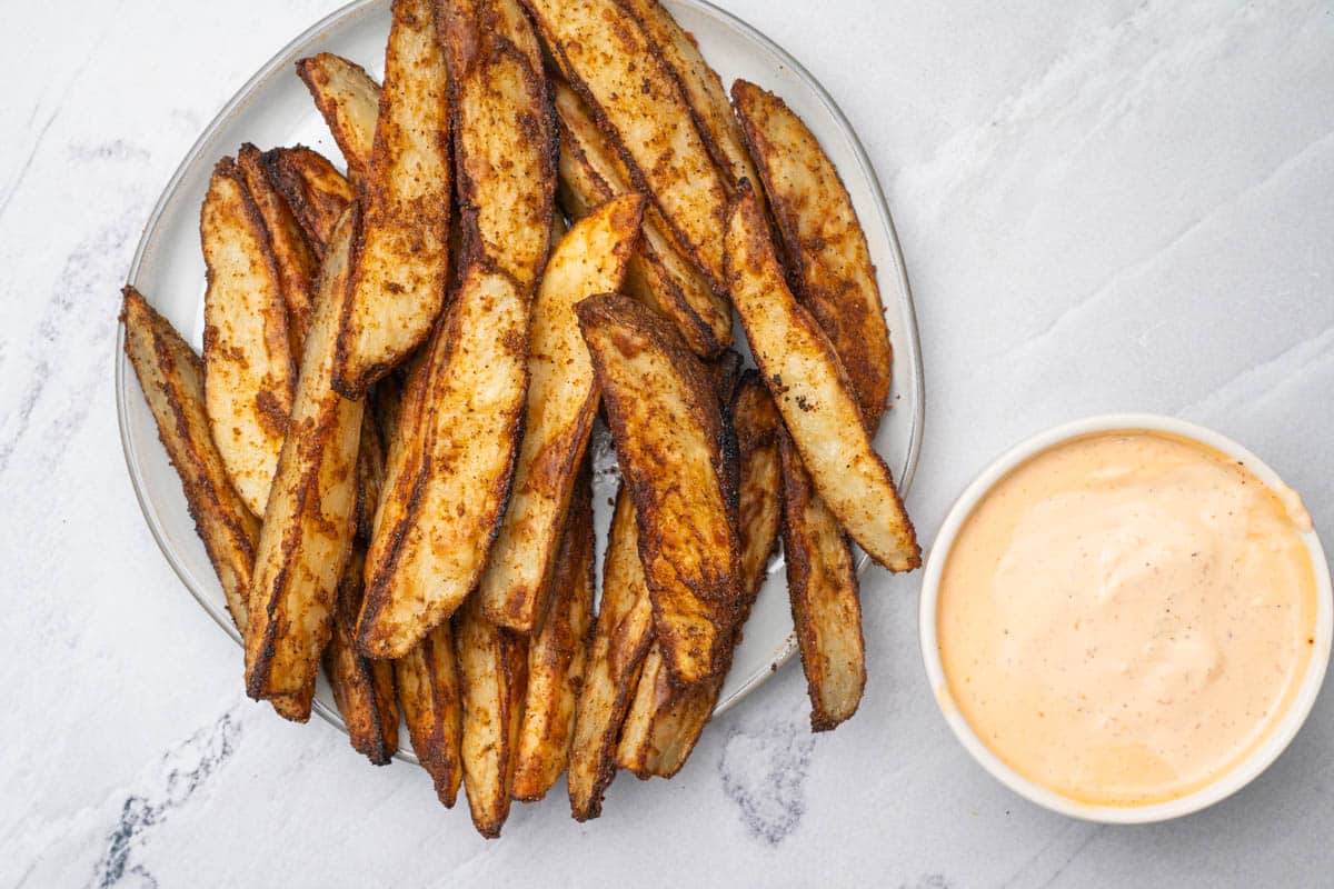 Oven roasted potato wedges on a round plate with sriracha aioli in a white bowl.